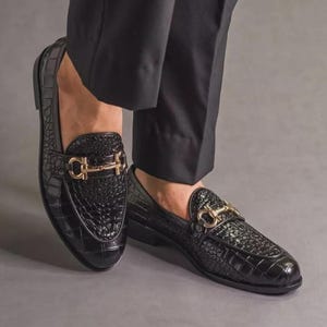 K&ouml;nnte beinhalten: Schwarze Leder-Loafer mit Krokodiltextur und goldfarbener Hardware. Die Schuhe haben ein klassisches Design mit einem Slip-on-Stil. Das Bild zeigt die Schuhe aus einem niedrigen Winkel, wobei die Textur und die Details hervorgehoben werden.