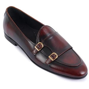 May include: Burgundy leather double monk strap shoes with gold-colored buckles. The shoes have a classic design with a rounded toe and a smooth, polished finish. The leather has a rich, deep color.