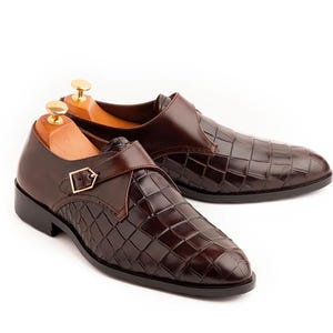 Pode incluir: Um par de sapatos de couro castanho com um padr&atilde;o texturizado semelhante ao de um crocodilo. Cada sapato apresenta uma tira com uma fivela dourada. Os sapatos s&atilde;o exibidos em formas de madeira sobre um fundo branco.