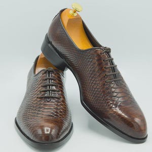 May include: A pair of brown leather dress shoes with a textured, snakeskin-like pattern. The shoes have a classic lace-up design, a black sole, and are displayed with wooden shoe trees inside. The shoes are a rich, dark brown color.