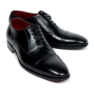 May include: A pair of black leather dress shoes with a glossy finish. The shoes have a classic cap-toe design, black laces, and red interior lining. The shoes are displayed on a white background.