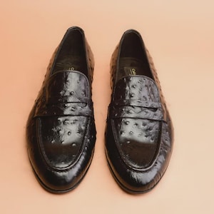 May include: A pair of black leather penny loafers with a textured, dotted pattern. The shoes have a classic design with a strap across the vamp and a rounded toe. The loafers are displayed on a light brown surface.