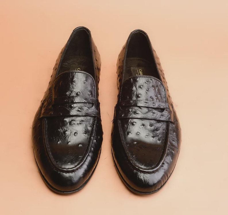 May include: A pair of black leather penny loafers with a textured, dotted pattern. The shoes have a classic design with a strap across the vamp and a rounded toe. The loafers are displayed on a light brown surface.
