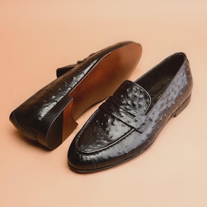 May include: Pair of black leather loafers with a textured surface, featuring a slip-on design. The shoes have a glossy finish and a brown sole. The background is a solid, light peach color.