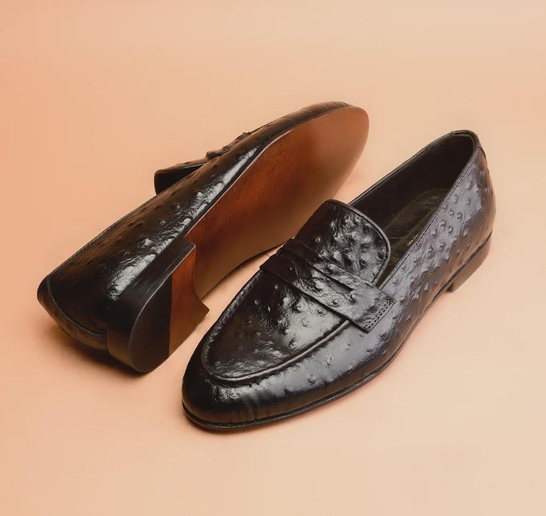 May include: Pair of black leather loafers with a textured surface, featuring a slip-on design. The shoes have a glossy finish and a brown sole. The background is a solid, light peach color.