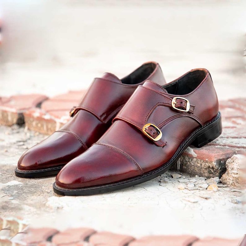 Peut inclure: Paire de chaussures &agrave; double boucle en cuir bordeaux. Elles pr&eacute;sentent une finition polie, des boucles dor&eacute;es et une semelle noire. Les chaussures sont pos&eacute;es sur une surface en briques, mettant en valeur leur design formel et leur savoir-faire.