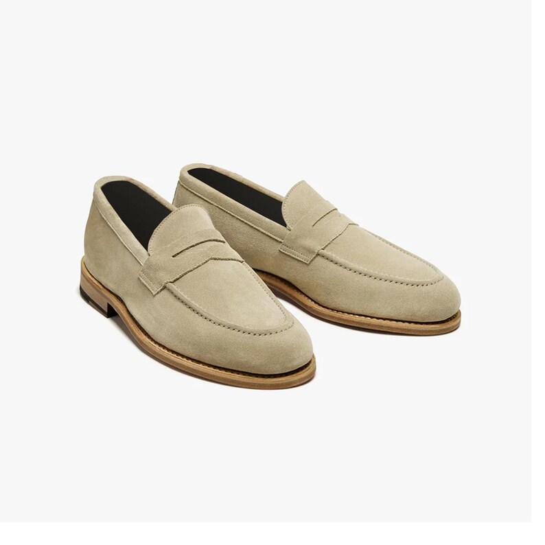 May include: A pair of beige suede penny loafers. The shoes have a classic design with a rounded toe, a strap across the vamp, and a stacked leather sole. The interior is black.