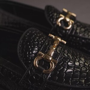 K&ouml;nnte beinhalten: Nahaufnahme von schwarzen Leder-Loafern mit Krokodiltextur. Die Schuhe haben goldfarbene Metallbeschl&auml;ge an den Riemen. Die Loafer sind ein klassischer Stil, geeignet f&uuml;r formelle oder l&auml;ssige Kleidung.