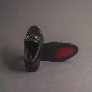 K&ouml;nnte beinhalten: Schwarze Leder-Loafer mit Krokodiltextur. Die Schuhe haben eine goldfarbene Schnalle und eine rote Sohle. Das Wort "FEATH" ist auf der Seite sichtbar. Diese stilvollen Loafer sind eine klassische Schuhwahl.
