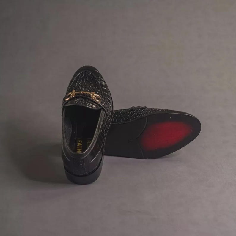 K&ouml;nnte beinhalten: Schwarze Leder-Loafer mit Krokodiltextur. Die Schuhe haben eine goldfarbene Schnalle und eine rote Sohle. Das Wort "FEATH" ist auf der Seite sichtbar. Diese stilvollen Loafer sind eine klassische Schuhwahl.