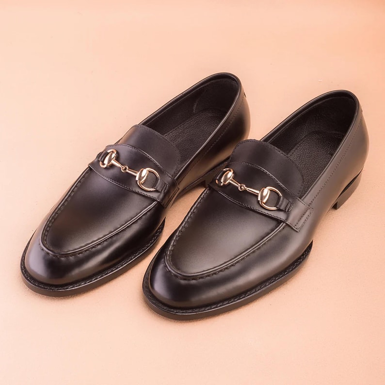 K&ouml;nnte beinhalten: Ein Paar schwarze Leder-Loafer mit einem goldfarbenen Metall-Bit-Detail. Die Schuhe haben ein klassisches Slip-on-Design mit runder Spitze und kleinem Absatz. Die Loafer werden auf einer hellbraunen Oberfl&auml;che pr&auml;sentiert.