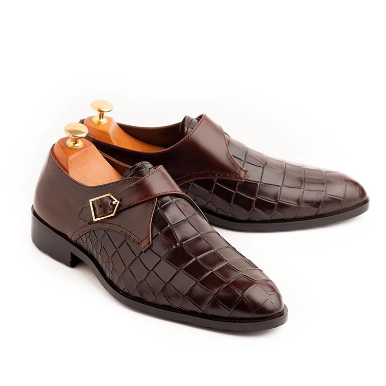 Pode incluir: Um par de sapatos de couro castanho com um padr&atilde;o texturizado semelhante ao de um crocodilo. Cada sapato apresenta uma tira com uma fivela dourada. Os sapatos s&atilde;o exibidos em formas de madeira sobre um fundo branco.