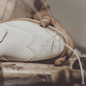 May include: Close-up of a white leather shoe being hand-stitched. The shoe features a buckle closure and decorative perforations. The shoemaking process is highlighted, showcasing craftsmanship and attention to detail in footwear.