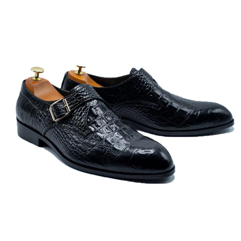 May include: A pair of black leather dress shoes with a crocodile texture. The shoes have a single strap with a gold-coloured buckle. Wooden shoe trees are inside the shoes.