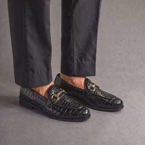K&ouml;nnte beinhalten: Schwarze Leder-Loafer mit Krokodiltextur und goldfarbener Hardware. Die Schuhe werden mit einer schwarzen Hose getragen. Die Loafer haben ein klassisches Design, das f&uuml;r formelle oder halbformelle Anl&auml;sse geeignet ist.