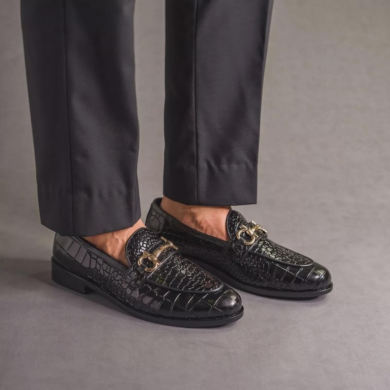 K&ouml;nnte beinhalten: Schwarze Leder-Loafer mit Krokodiltextur und goldfarbener Hardware. Die Schuhe werden mit einer schwarzen Hose getragen. Die Loafer haben ein klassisches Design, das f&uuml;r formelle oder halbformelle Anl&auml;sse geeignet ist.