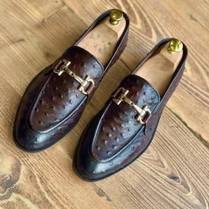 May include: A pair of brown leather loafers with a textured finish and gold-coloured metal buckle detail. The shoes have a classic design and are displayed on a wooden surface. Shoe trees are inside the shoes.