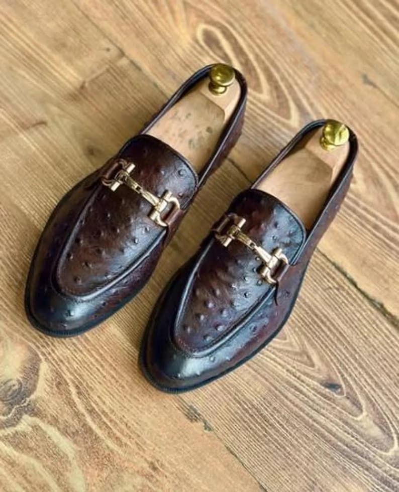 May include: A pair of brown leather loafers with a textured finish and gold-coloured metal buckle detail. The shoes have a classic design and are displayed on a wooden surface. Shoe trees are inside the shoes.