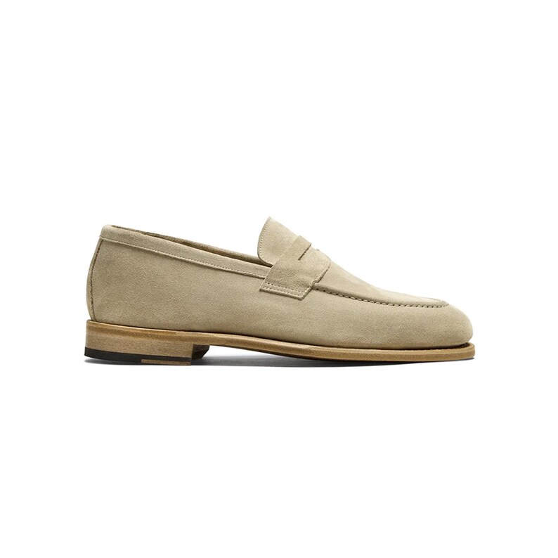 May include: A beige suede penny loafer shoe. The shoe features a rounded toe, a strap across the vamp, and a low heel. The sole is a light brown color, and the shoe has a classic, minimalist design.