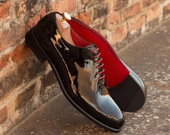 May include: A pair of black patent leather dress shoes with red laces and a red interior. The shoes are leaning against a brick wall. The sole of the shoe is black with a gray stripe. A wooden shoe tree is inside one shoe.