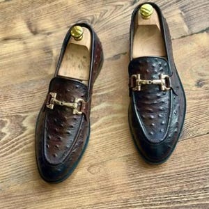 May include: A pair of brown leather loafers with a textured finish and gold-coloured metal accents. The shoes have a classic design with a slip-on style and are displayed on a wooden surface.