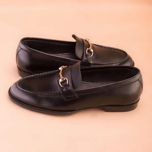 K&ouml;nnte beinhalten: Ein Paar schwarze Leder-Loafer mit goldfarbenen Metallakzenten. Die Schuhe haben ein klassisches Design mit runder Spitze und einem Slip-on-Stil. Das goldfarbene Metalldetail befindet sich auf der Oberseite des Schuhs.