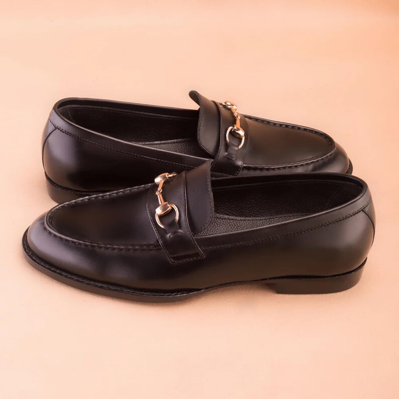 K&ouml;nnte beinhalten: Ein Paar schwarze Leder-Loafer mit goldfarbenen Metallakzenten. Die Schuhe haben ein klassisches Design mit runder Spitze und einem Slip-on-Stil. Das goldfarbene Metalldetail befindet sich auf der Oberseite des Schuhs.