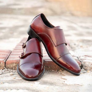 Peut inclure: Une paire de chaussures en cuir bordeaux &agrave; boucle de moine avec des boucles dor&eacute;es. Les chaussures ont une finition polie et sont pr&eacute;sent&eacute;es sur une surface en briques. Les chaussures ont un design classique.