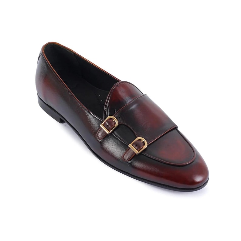May include: Burgundy leather double monk strap shoes with gold-colored buckles. The shoes have a classic design with a rounded toe and a smooth, polished finish. The leather has a rich, deep color.