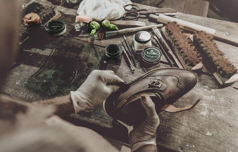 May include: A brown leather shoe with brogue detailing is being worked on. Shoe care tools, including brushes and creams, are arranged on a wooden surface. The scene suggests shoe repair or restoration, with the person wearing gloves.