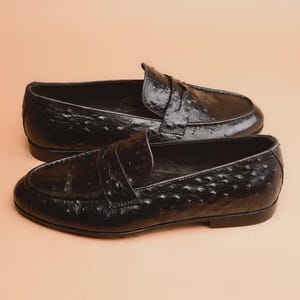 May include: A pair of black leather penny loafers with a textured, patterned surface. The shoes have a classic design with a strap across the top and a low heel. The loafers are displayed on a neutral background.
