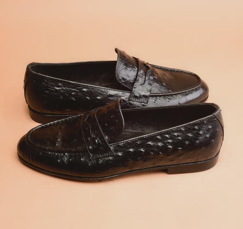 May include: A pair of black leather penny loafers with a textured, patterned surface. The shoes have a classic design with a strap across the top and a low heel. The loafers are displayed on a neutral background.