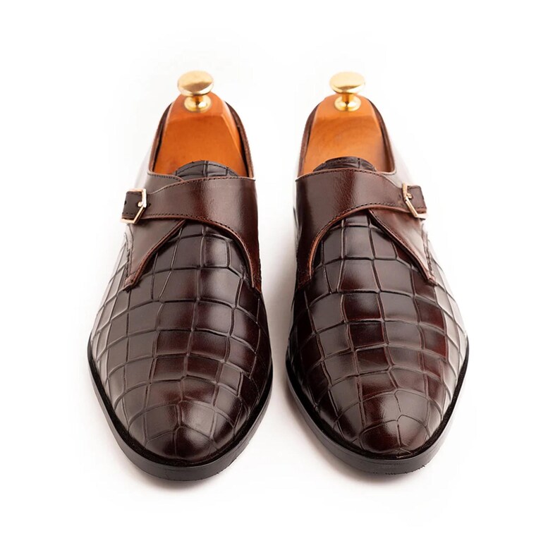Pode incluir: Um par de sapatos de couro castanho escuro com um padr&atilde;o texturizado xadrez. Os sapatos apresentam uma tira com fecho de fivela e s&atilde;o exibidos em formas de madeira. Os sapatos s&atilde;o de cor castanho escuro.