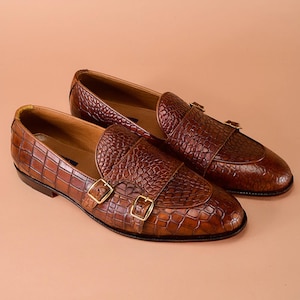 Puede incluir: Un par de mocasines de cuero marrón con textura de piel de cocodrilo. Los zapatos presentan correas dobles con hebillas doradas. Los mocasines se exhiben sobre una superficie marrón claro.