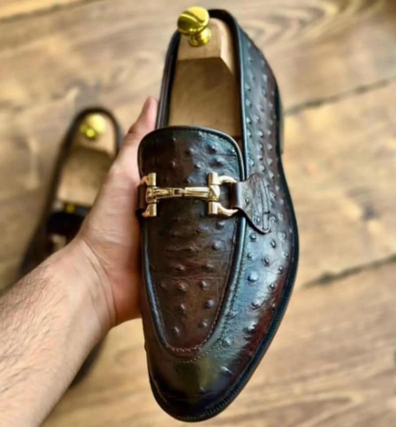May include: A brown leather loafer with a gold buckle. The shoe has a textured surface and a classic design. Another shoe is visible in the background, out of focus.