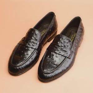 May include: A pair of black leather loafers with a textured finish. The shoes have a classic slip-on design with a strap across the top. The shoes are displayed on a light peach-colored surface.