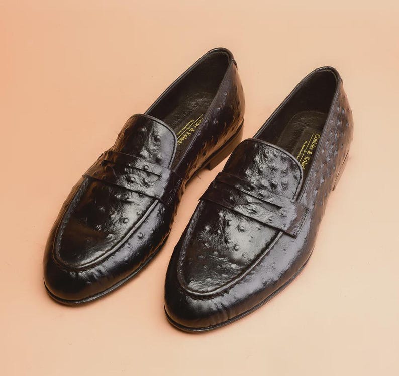 May include: A pair of black leather loafers with a textured finish. The shoes have a classic slip-on design with a strap across the top. The shoes are displayed on a light peach-colored surface.