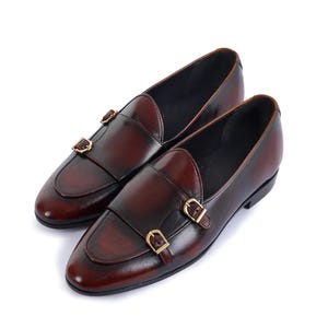 May include: A pair of burgundy leather loafers with double monk straps and gold-colored buckles. The shoes have a classic design with a rounded toe and a low heel. The leather has a polished finish.