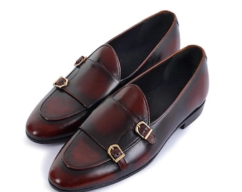 Può includere: Un paio di mocassini in pelle bordeaux con doppi cinturini e fibbie dorate. Queste scarpe classiche hanno una punta arrotondata e un tacco basso. La pelle ha una finitura lucida. Il colore è rosso scuro.