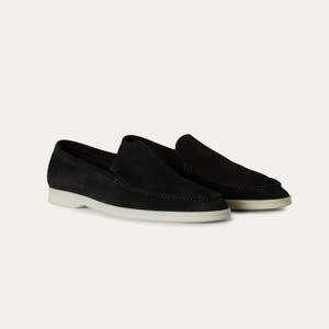 K&ouml;nnte beinhalten: Ein Paar schwarze Wildleder-Loafer mit wei&szlig;er Sohle. Die Schuhe haben ein klassisches Slip-on-Design und sind aus einem weichen, strukturierten Material gefertigt. Die Schuhe werden auf wei&szlig;em Hintergrund pr&auml;sentiert.
