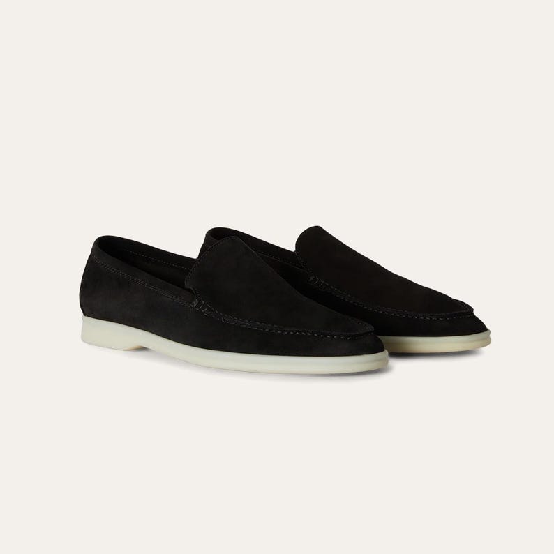 K&ouml;nnte beinhalten: Ein Paar schwarze Wildleder-Loafer mit wei&szlig;er Sohle. Die Schuhe haben ein klassisches Slip-on-Design und sind aus einem weichen, strukturierten Material gefertigt. Die Schuhe werden auf wei&szlig;em Hintergrund pr&auml;sentiert.