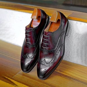 May include: Burgundy leather Oxford shoes with brogue detailing and wooden shoe trees. The shoes have a polished finish. A classic design suitable for formal wear. The shoes are displayed on a wooden surface.