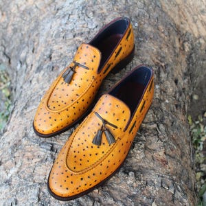 May include: A pair of yellow leather loafers with black dot detailing. Each shoe features a black tassel and a dark blue interior. The shoes are placed on a textured, brown surface.