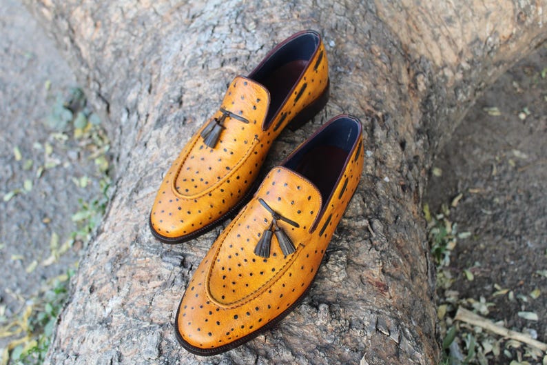 May include: A pair of yellow leather loafers with black dot detailing. Each shoe features a black tassel and a dark blue interior. The shoes are placed on a textured, brown surface.