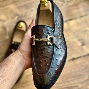 May include: A brown leather loafer with a gold-coloured buckle. The shoe has a textured surface and a polished finish. The shoe is held by a hand, with another shoe visible in the background. The shoe is a men's dress shoe.