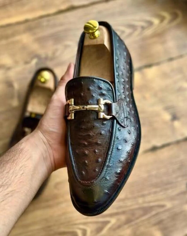 May include: A brown leather loafer with a gold-coloured buckle. The shoe has a textured surface and a polished finish. The shoe is held by a hand, with another shoe visible in the background. The shoe is a men's dress shoe.