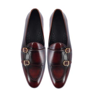 May include: A pair of burgundy leather dress shoes with double monk straps and gold-colored buckles. The shoes have a polished finish and a classic design, suitable for formal occasions.