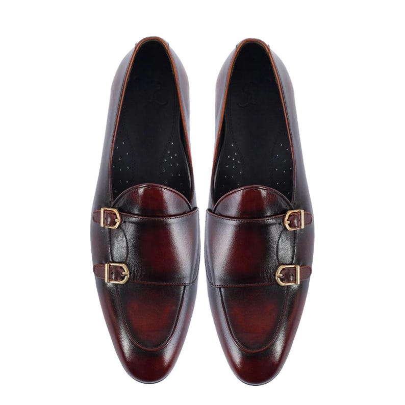 May include: A pair of burgundy leather dress shoes with double monk straps and gold-colored buckles. The shoes have a polished finish and a classic design, suitable for formal occasions.