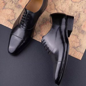 K&ouml;nnte beinhalten: Ein Paar schwarze Lederschuhe mit gl&auml;nzender Oberfl&auml;che. Die Schuhe haben ein klassisches Oxford-Design mit Schn&uuml;rverschluss und einer Kappe an der Spitze. Sie liegen auf einer Oberfl&auml;che mit Kartenmuster.