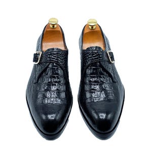 Könnte beinhalten: Ein Paar schwarze Lederschuhe mit Krokodilleder-Textur. Die Schuhe haben einen einzelnen Schnallenverschluss und werden auf hölzernen Schuhspannern präsentiert. Klassischer Stil.